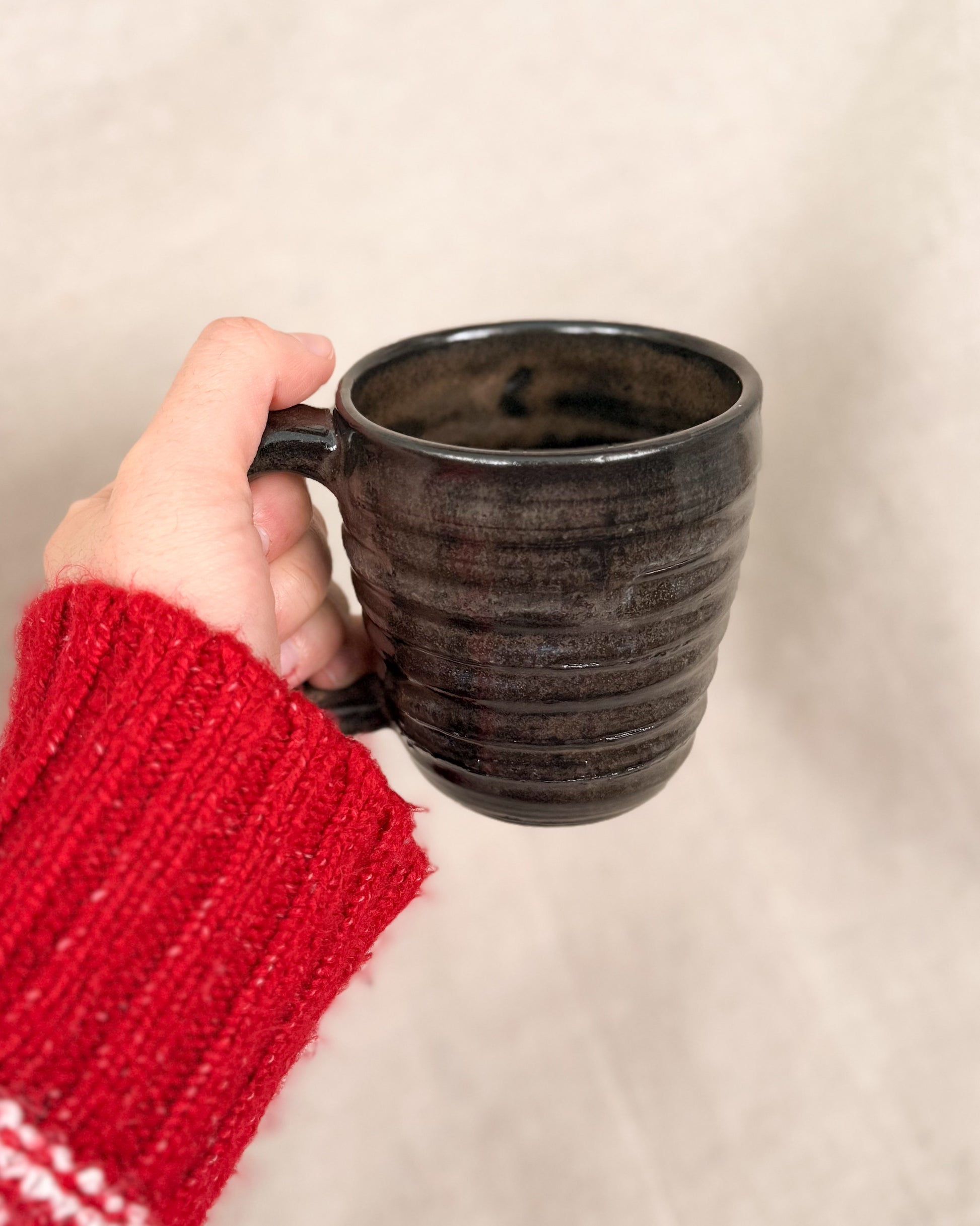 Hand holding a dark ceramic mug with a red sleeve against a beige background