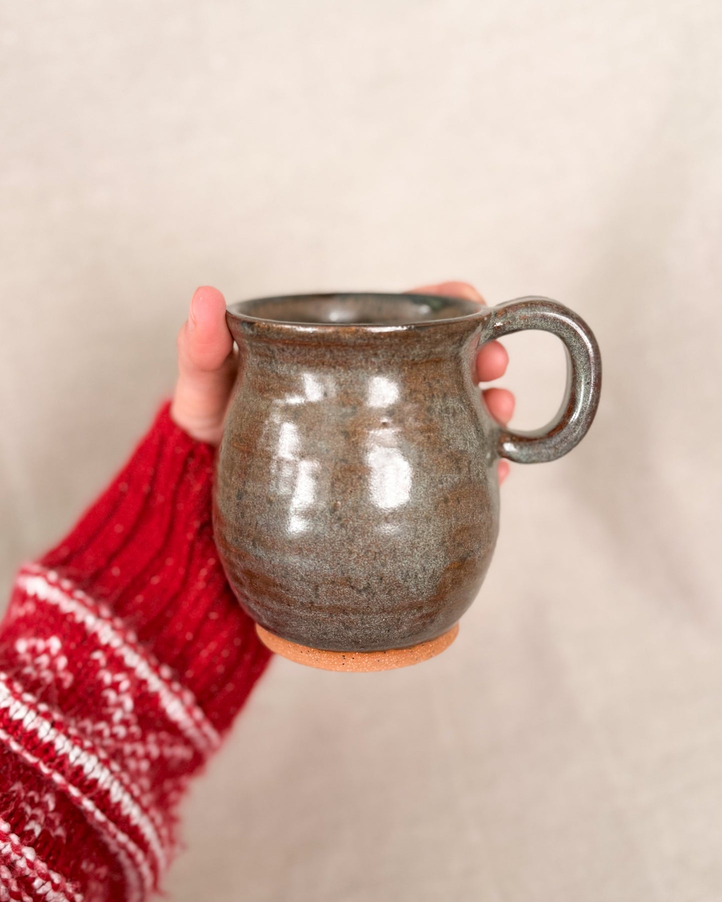 Hand holding a brown ceramic mug with a red and white patterned sleeve on a beige background