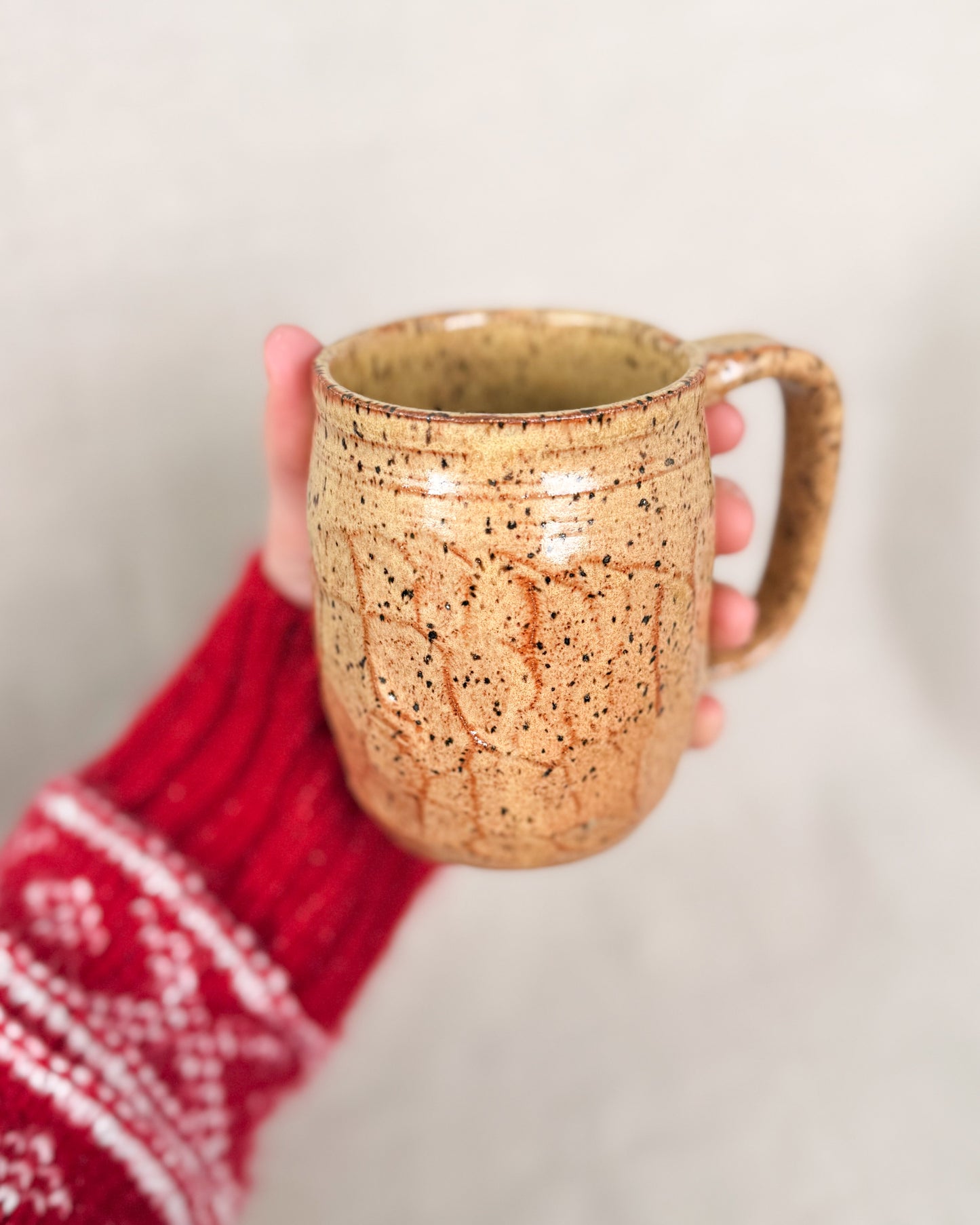 Hand holding a speckled ceramic mug with a red and white patterned sleeve on a light background