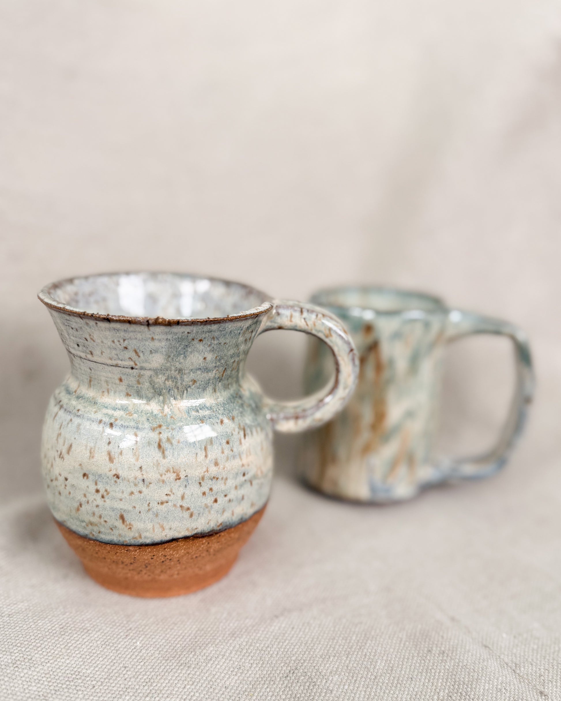 Two ceramic mugs with speckled glaze on a neutral background