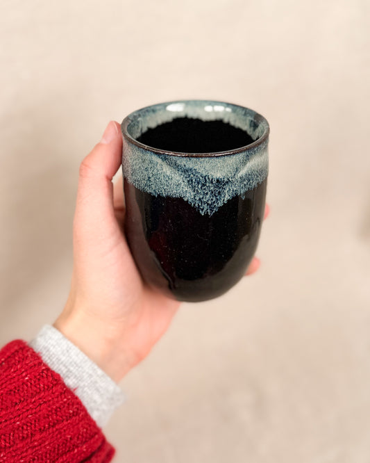 Hand holding a black ceramic mug with a textured surface against a beige background, 6 ounces