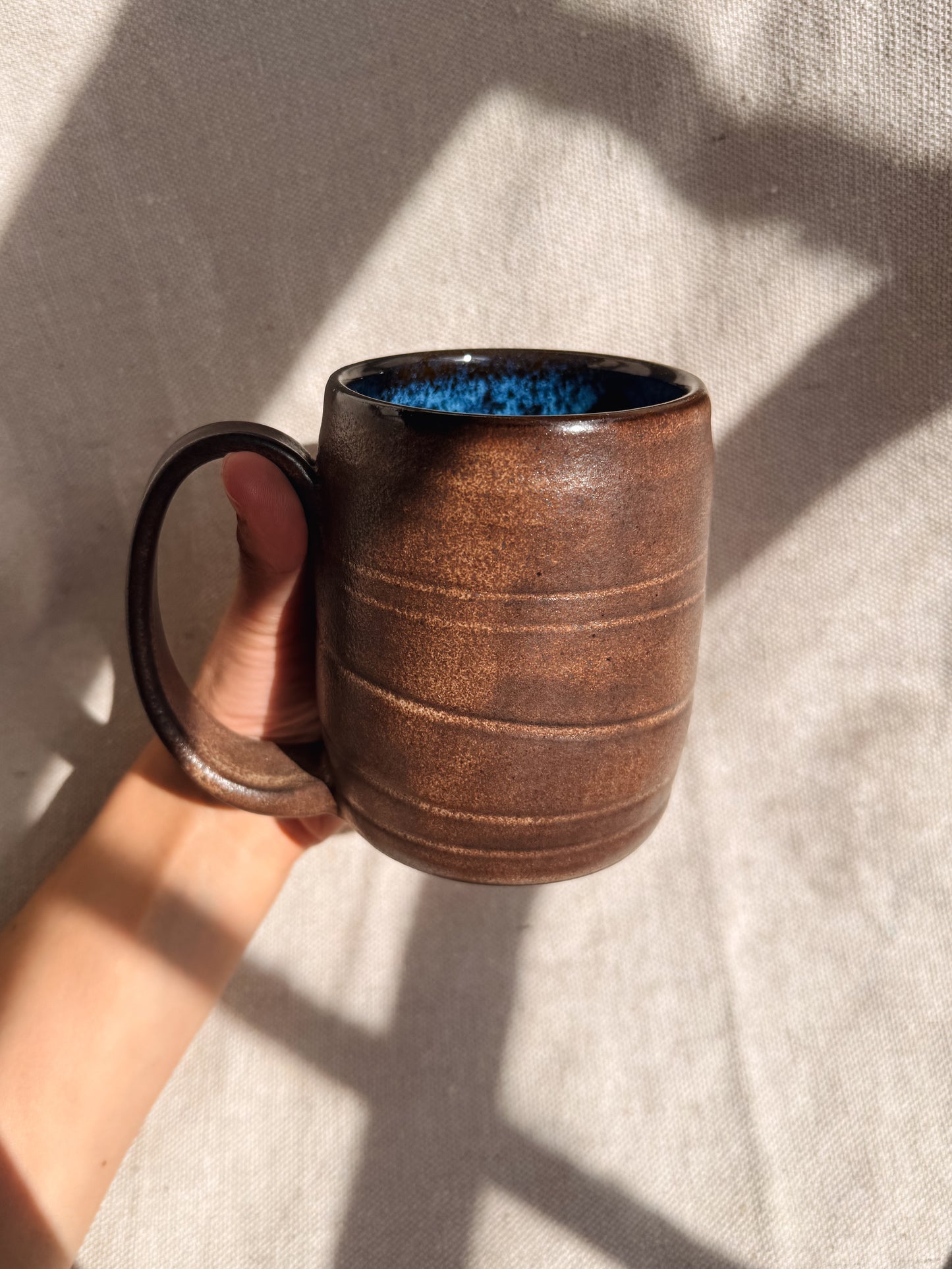 Brown ceramic mug with blue interior held by a hand against a neutral background