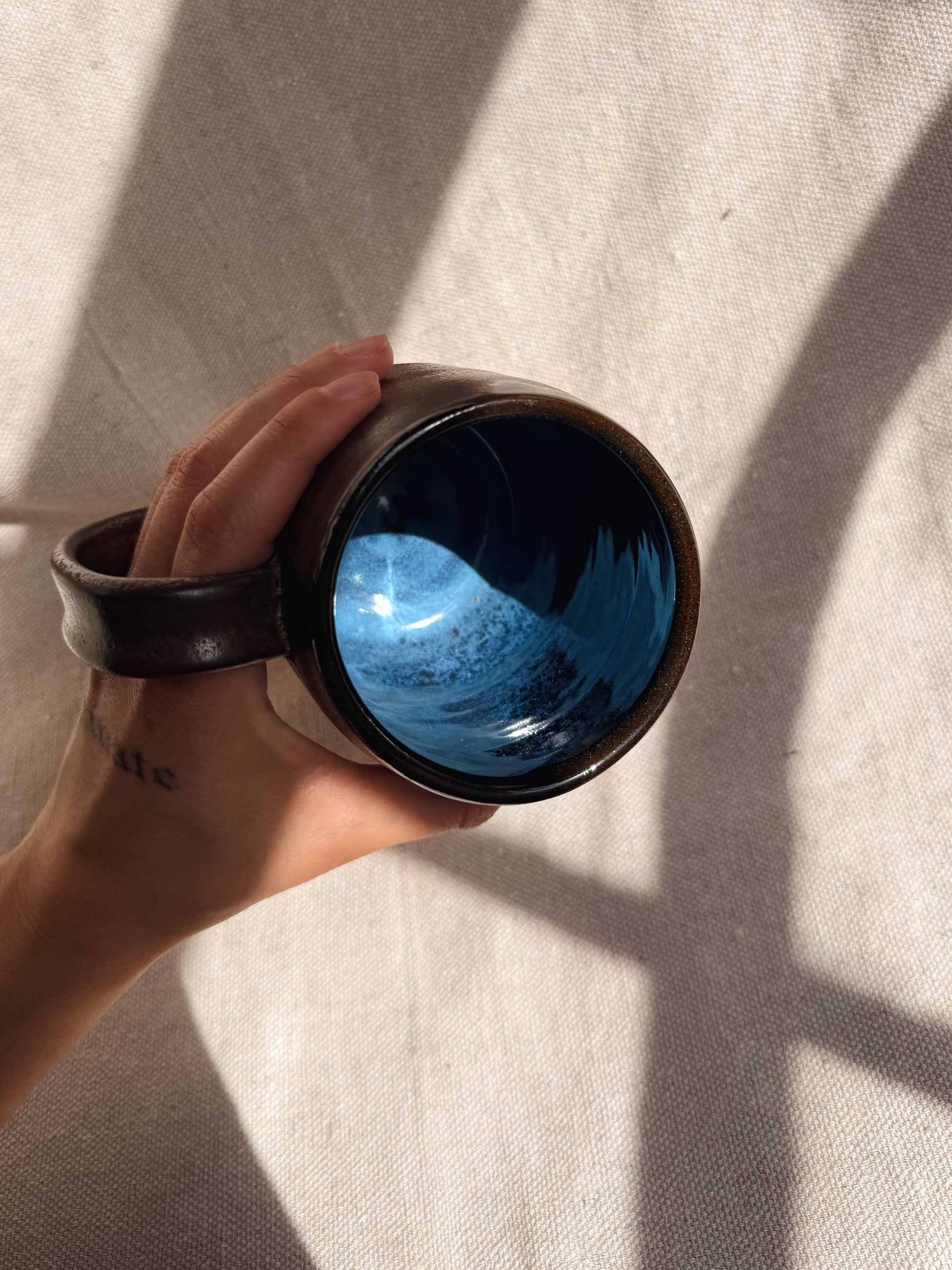 Hand holding a dark brown ceramic cup with blue interior against a light fabric background