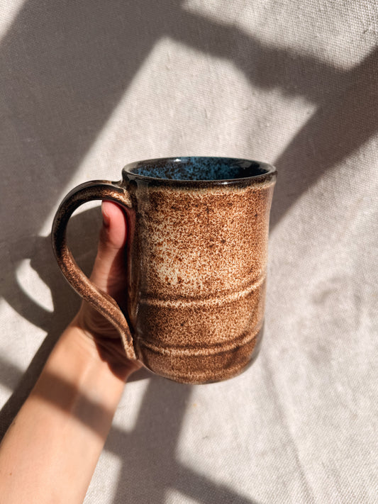 Hand holding a brown speckled ceramic mug with a blue interior against a light background