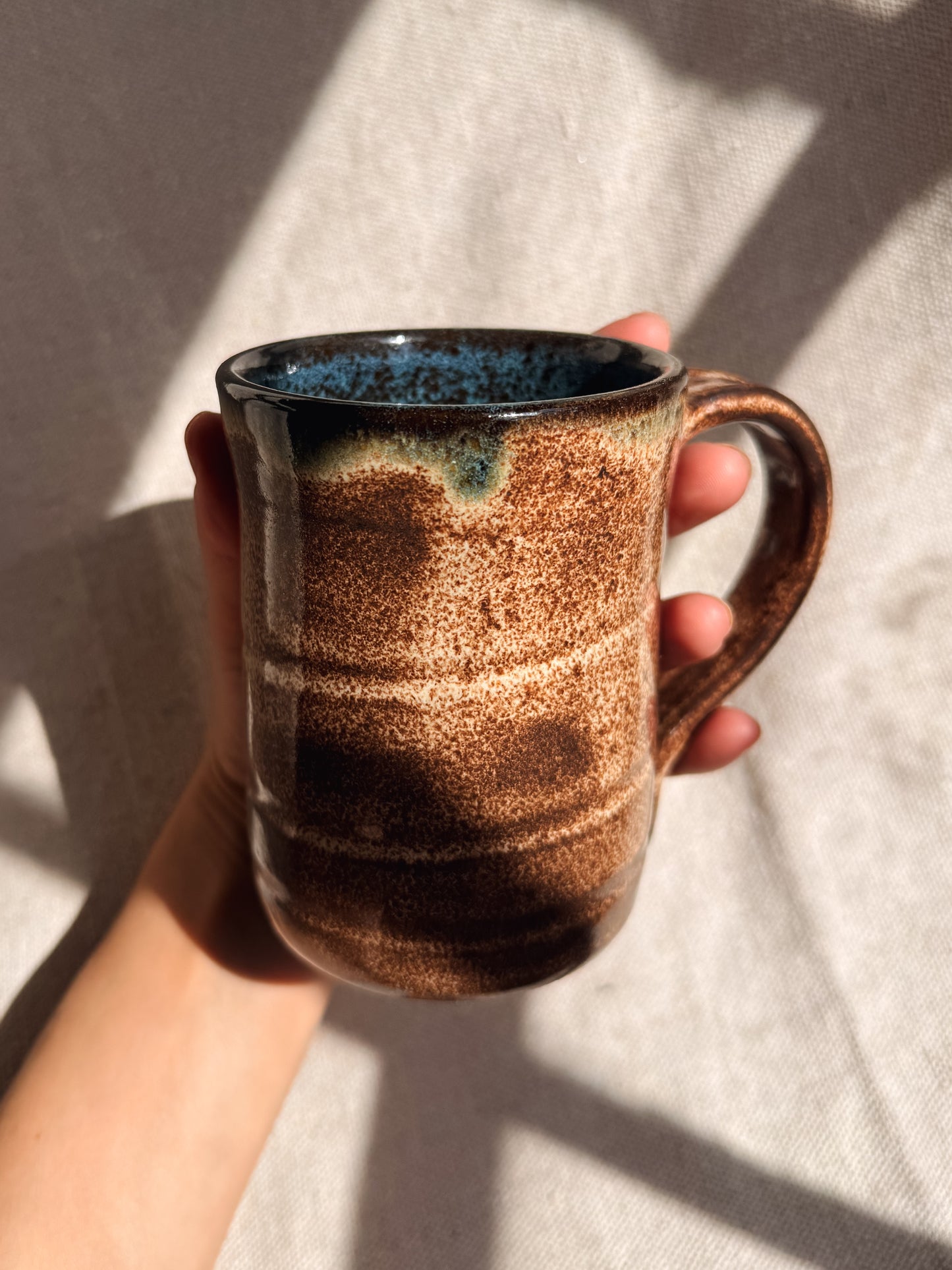 Hand holding a ceramic mug with brown and blue glaze against a neutral background