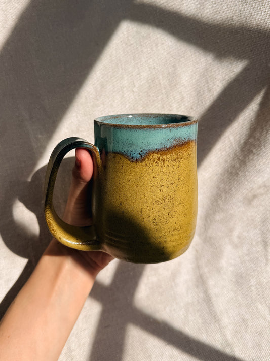 Hand holding a ceramic mug with a greenish-yellow body and blue top against a neutral background