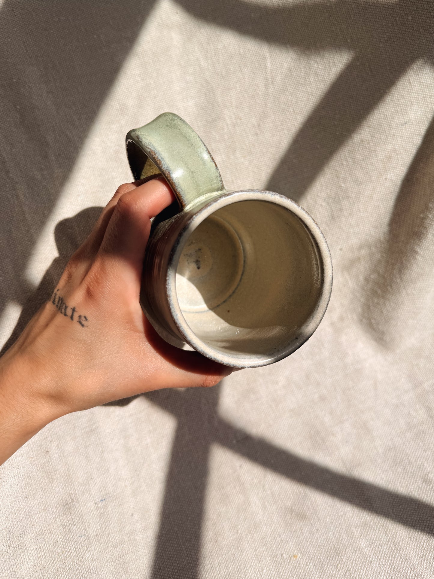 Hand holding a light green ceramic mug on a textured surface with shadows.