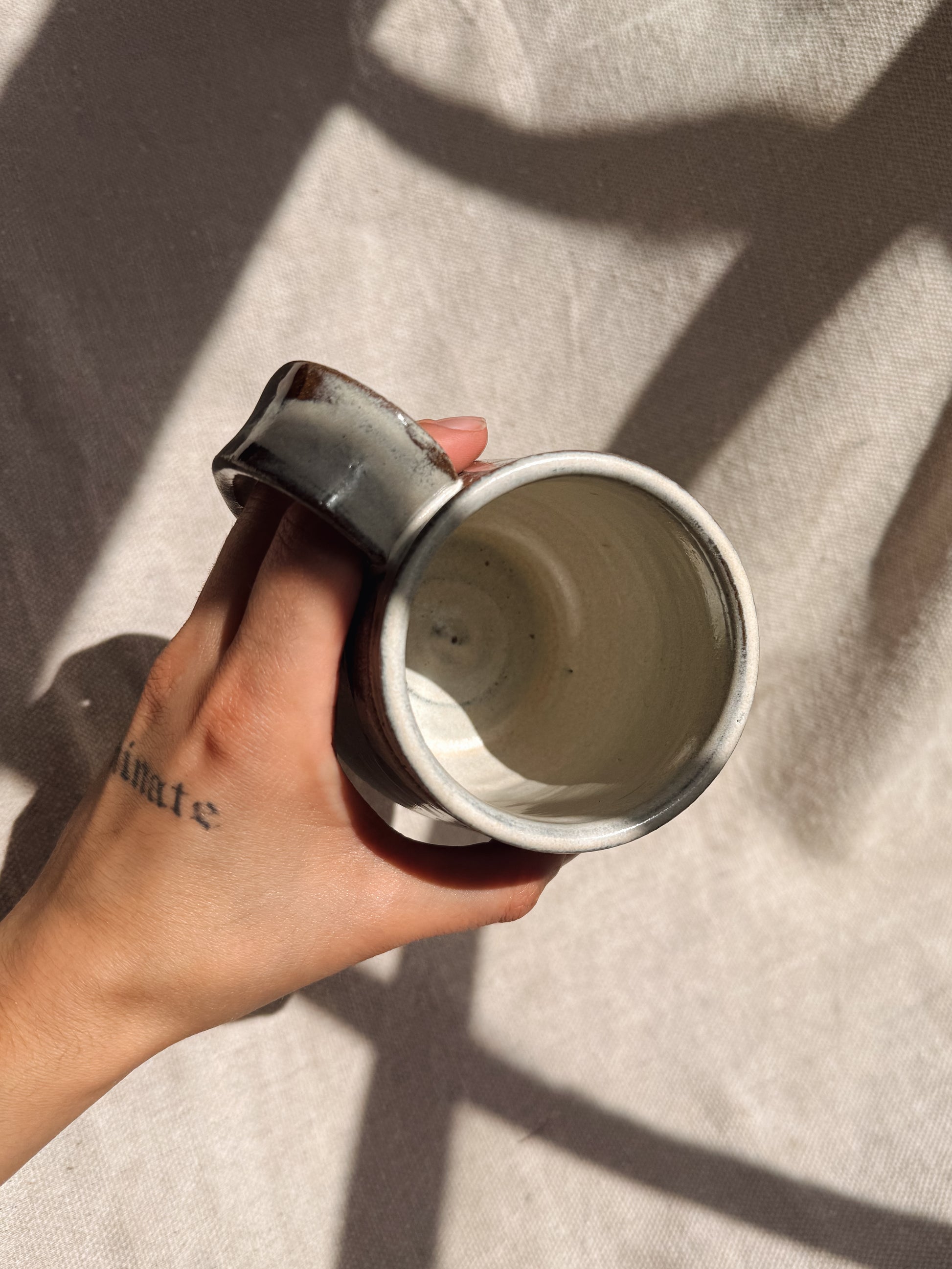Hand holding a ceramic mug with a tattoo on the arm against a textured beige background
