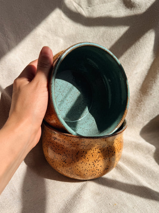 Hand holding a stack of ceramic bowls with a speckled brown exterior and teal interior on a beige fabric background.