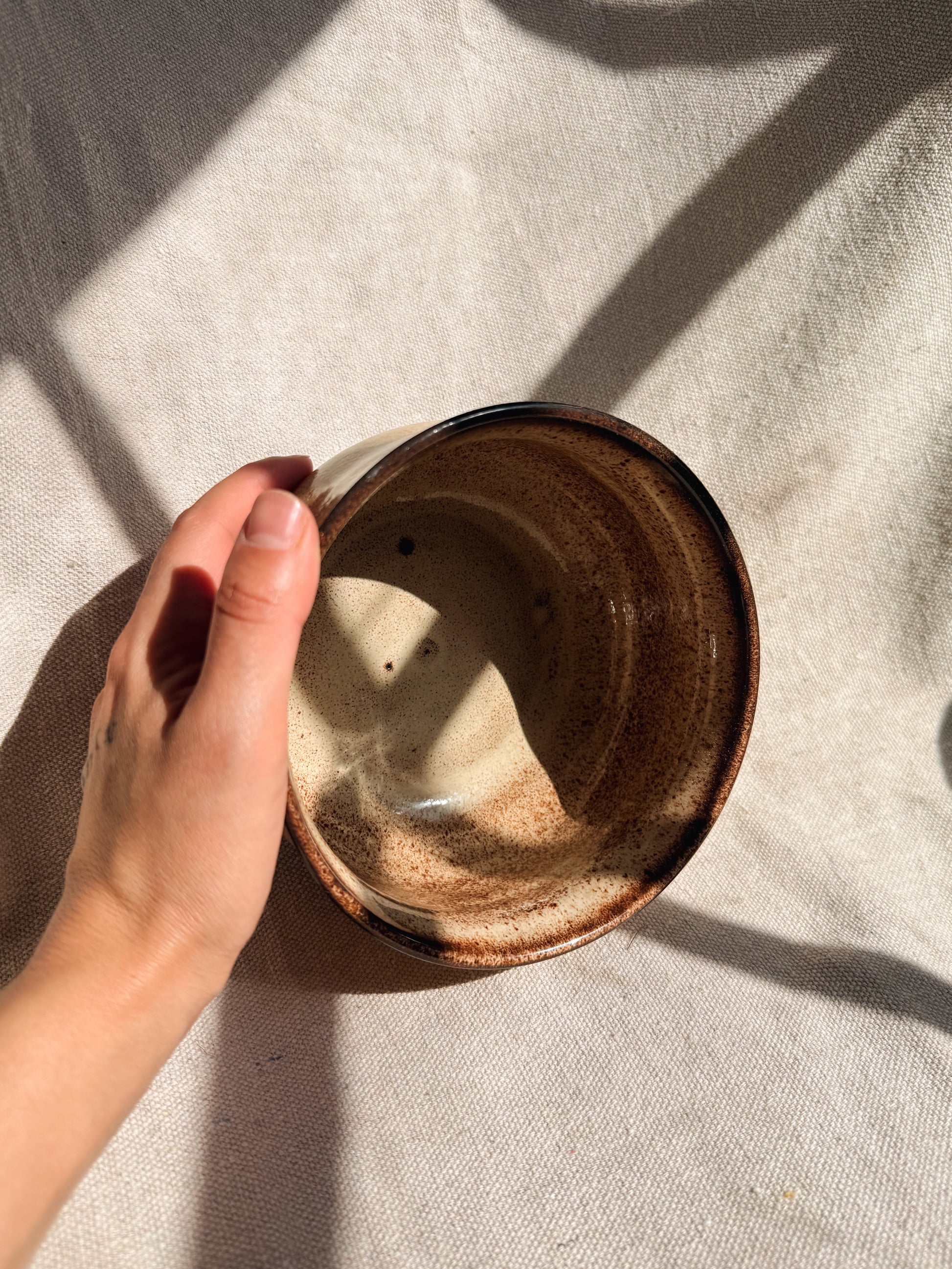 Hand holding a ceramic bowl with a brown glaze on a beige fabric background