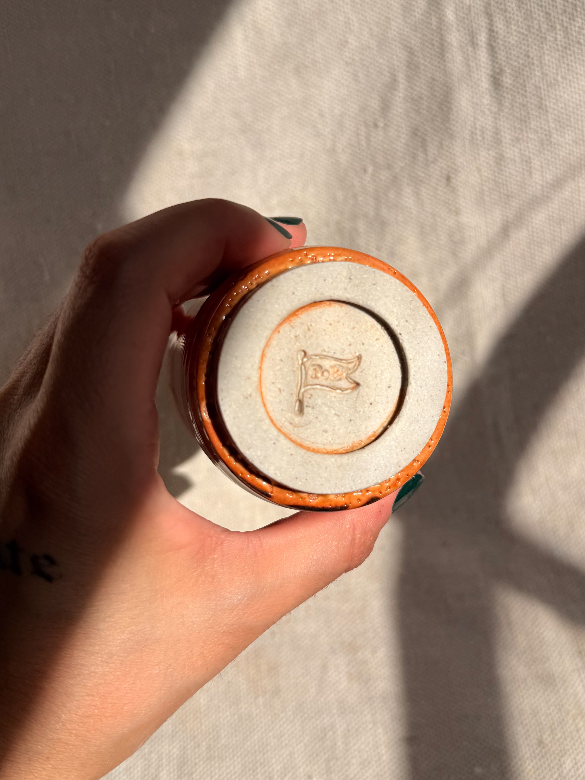 Hand holding a ceramic container with a logo on a neutral background