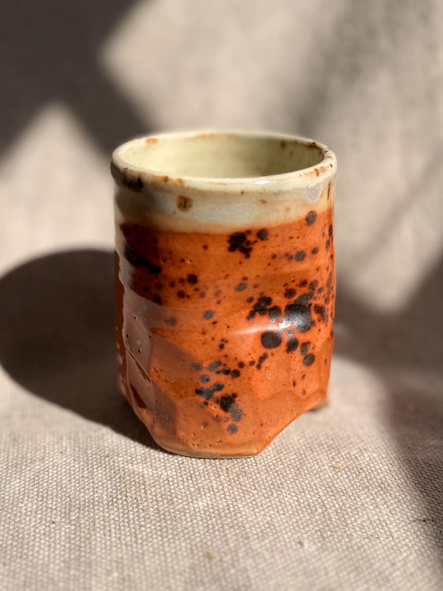 Small ceramic cup with orange glaze and black speckles on a textured surface
