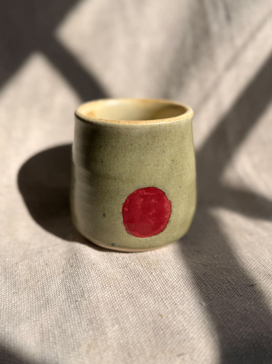 Green ceramic cup with a red dot on a textured beige surface