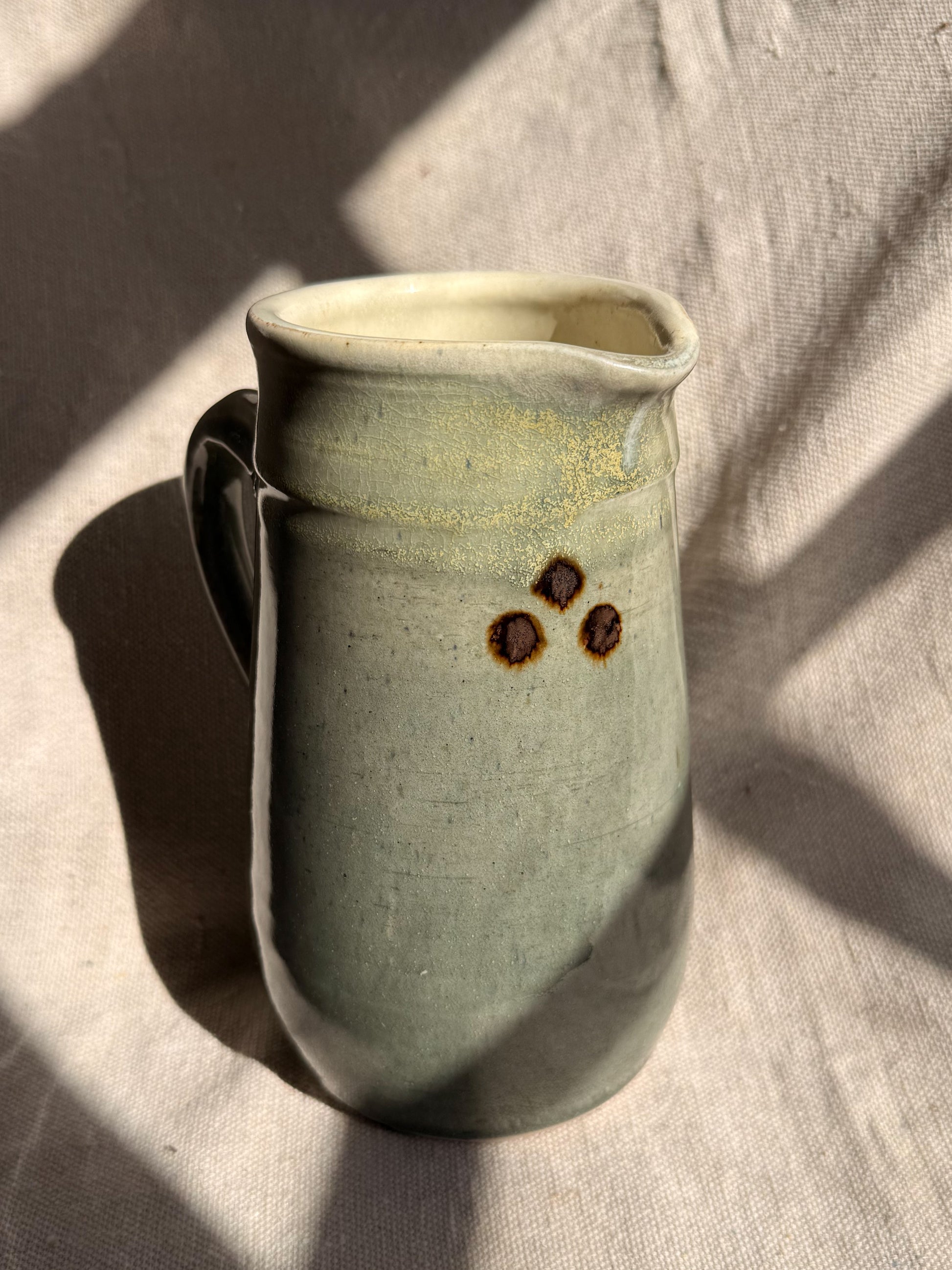 Green ceramic pitcher with brown spots on a textured surface